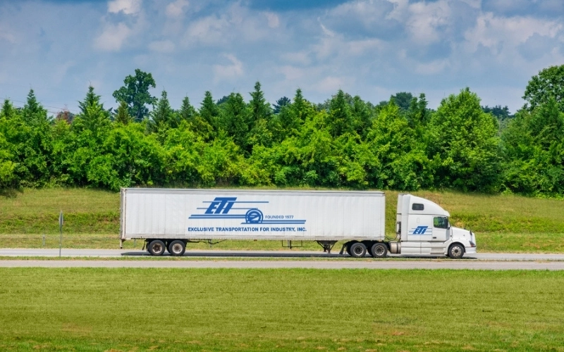 White tractor trailer with ETI logo on a road surrounded by grass, pine trees and clouds
